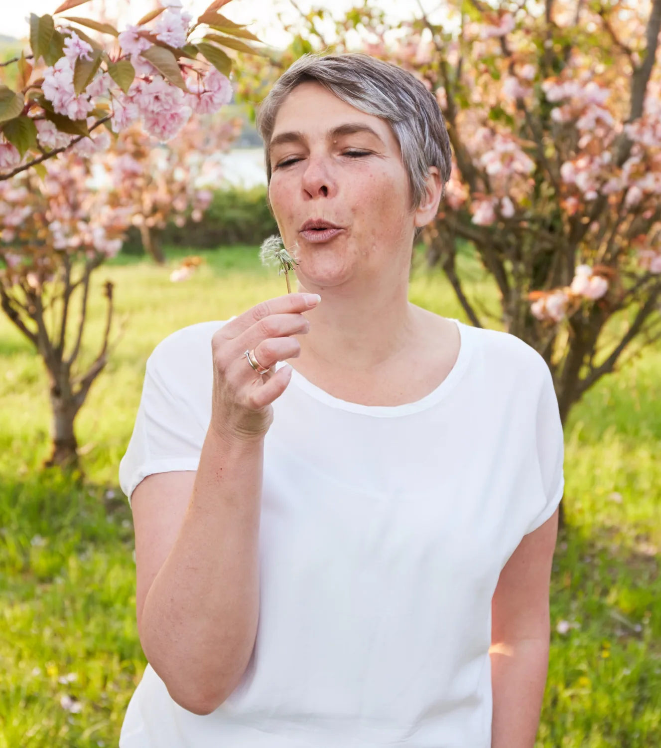 Janine Knüppel pustet lächelnd eine Pusteblume unter blühenden Kirschbäumen
